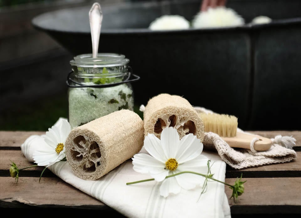 En person bag en krukke med hjemmelavet sukkerpeeling, blomster og en luffa.